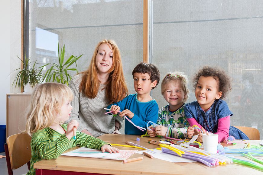 Kleinkinder sitzen mit ihrer Betreuerin am Tisch und basteln