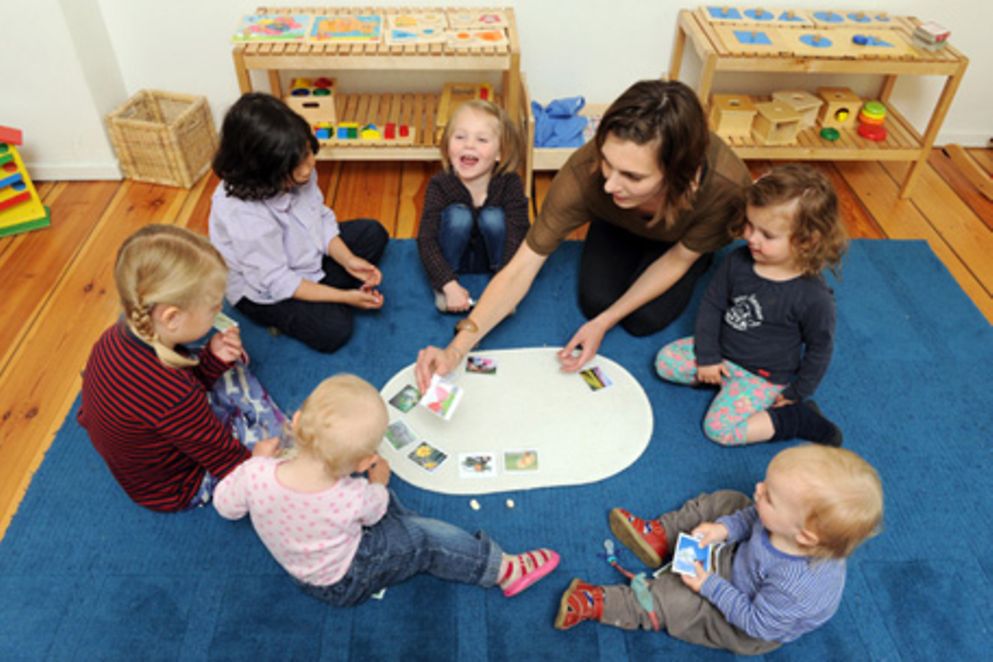 Mehrere Kinder und eine Betreuerin sitzen in einer Kita auf dem Boden und ordnen Karten zu.